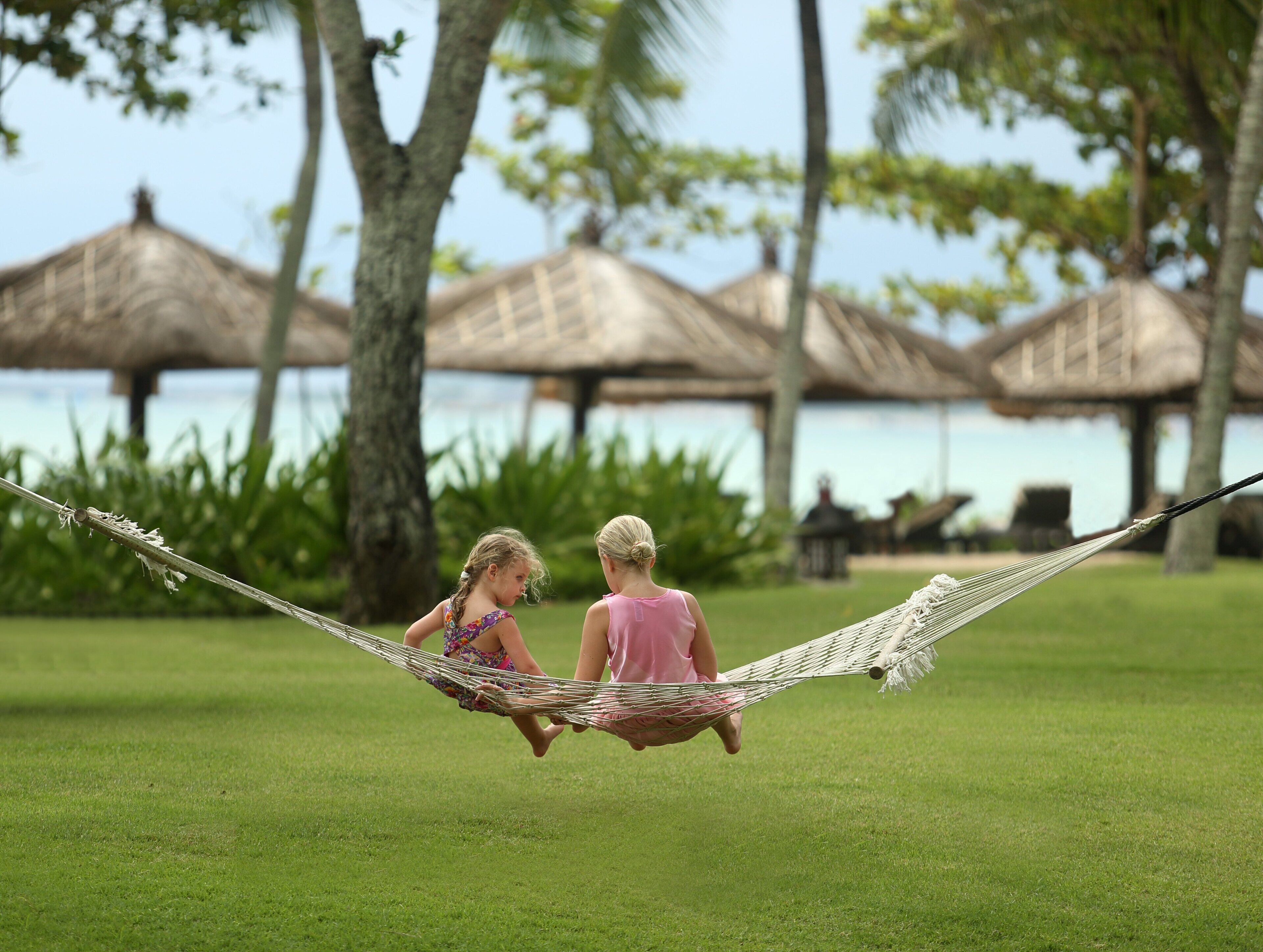 Two children swinging on a hammock in a grassy area with palm trees and thatched-roof structures in the background.
