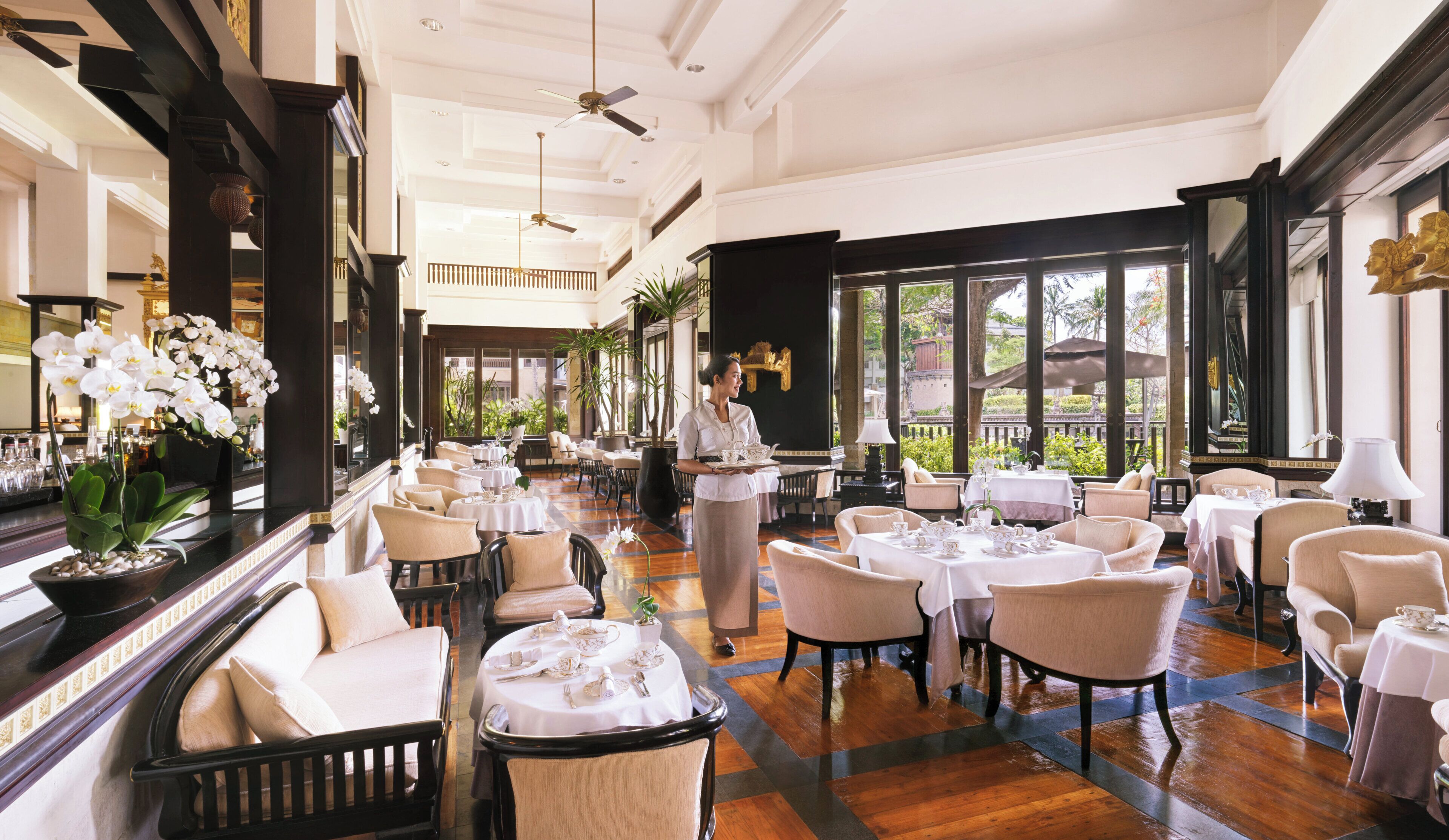A spacious hotel dining area with multiple tables and chairs, a waiter serving, and large windows offering views of the outdoors.