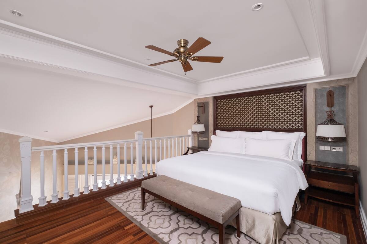 A bedroom with a large bed, a ceiling fan, a wooden bench, a nightstand with a lamp, and a patterned rug.