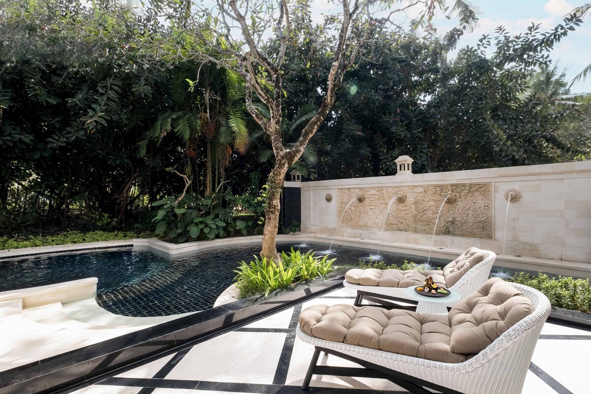 A pool area with a white wicker lounge chair, a small table with a decorative item, and a stone wall with a water feature.