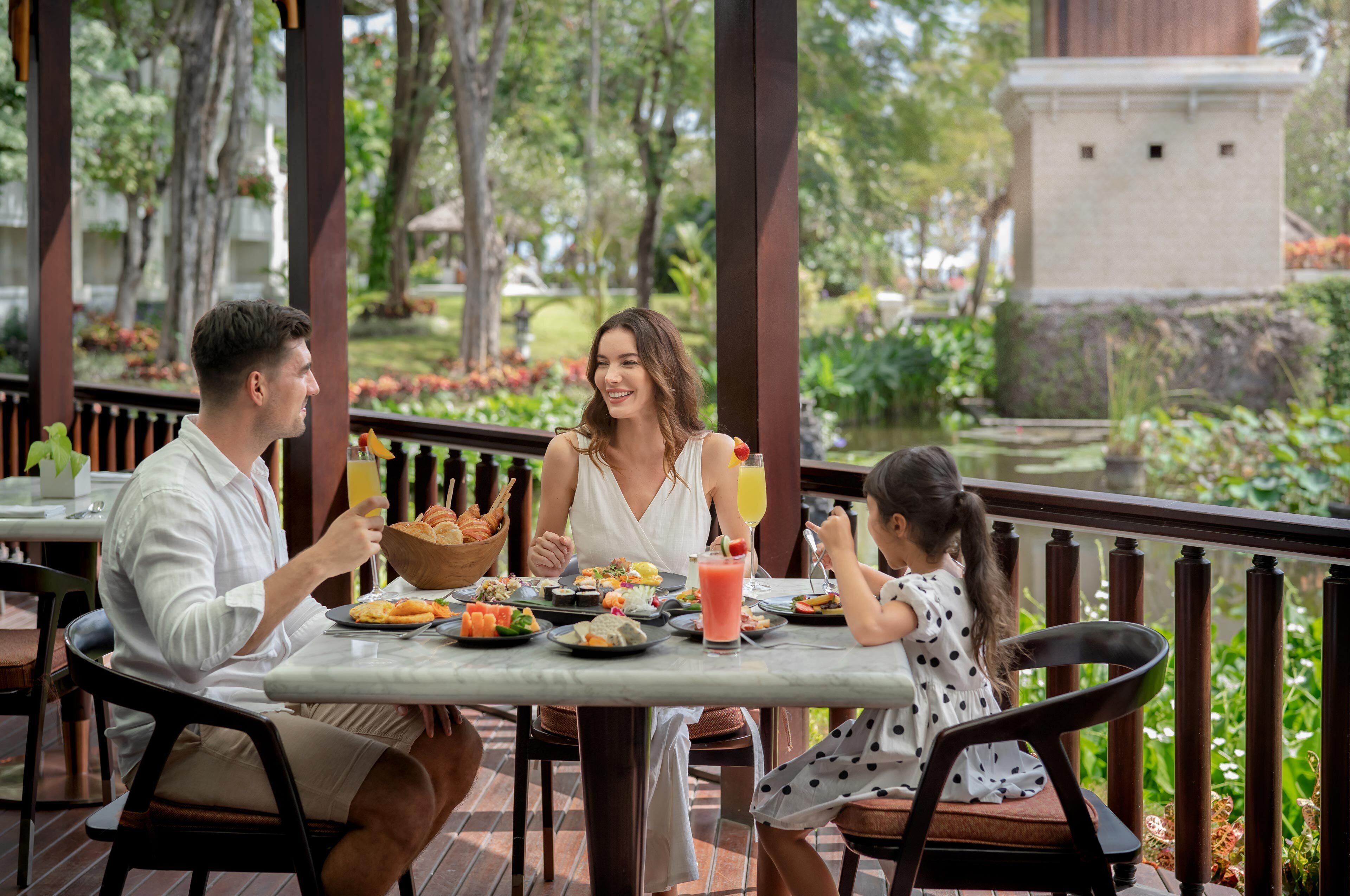A family dining at an outdoor table with a view of a garden and a statue.
