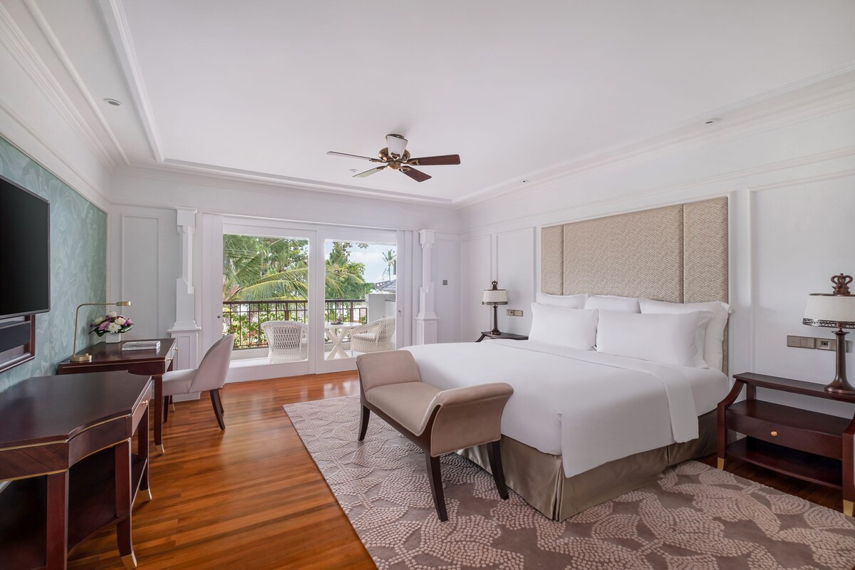 A hotel room with a large bed, a desk, a chair, a television, and a balcony with outdoor furniture.