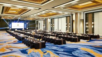 A large conference room with rows of tables, chairs, and floral centerpieces, featuring a blue and yellow carpet and a large screen at the front.