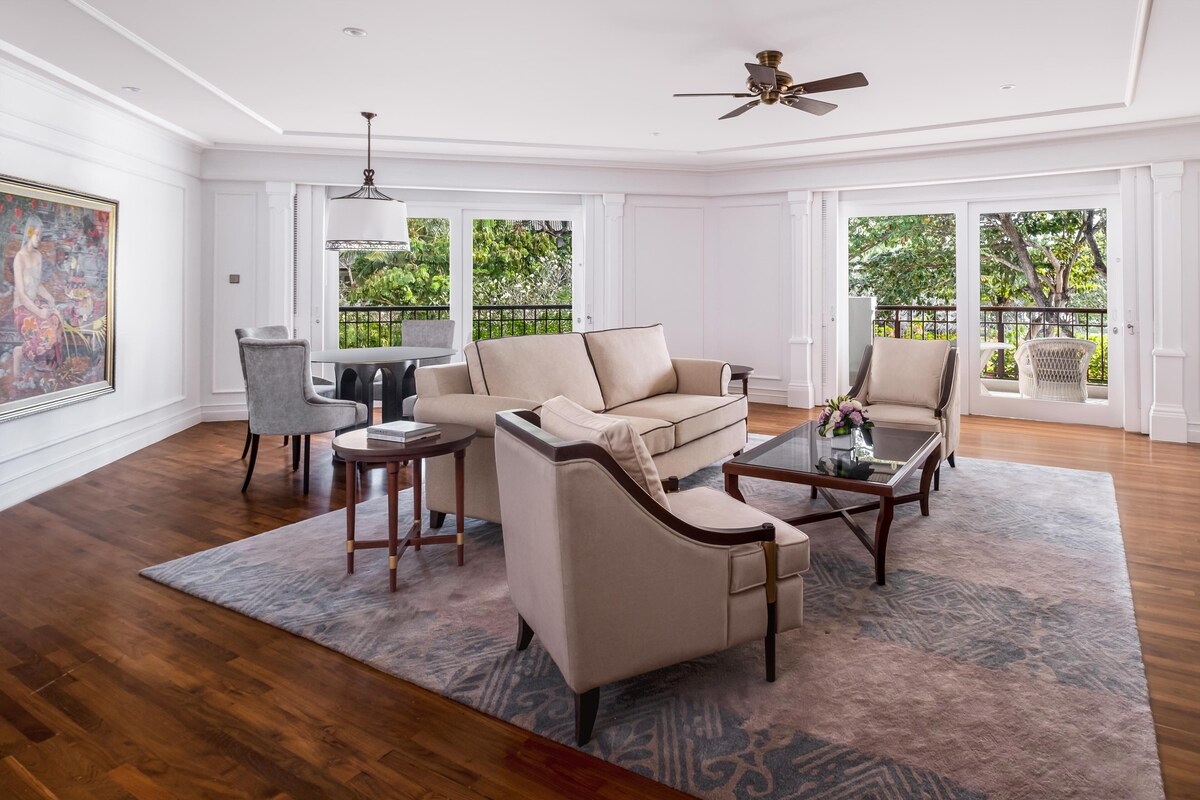 A spacious living room with a beige sofa, two armchairs, a coffee table, and a dining table. There is a painting on the wall, a ceiling fan, and large windows with a view of greenery outside.