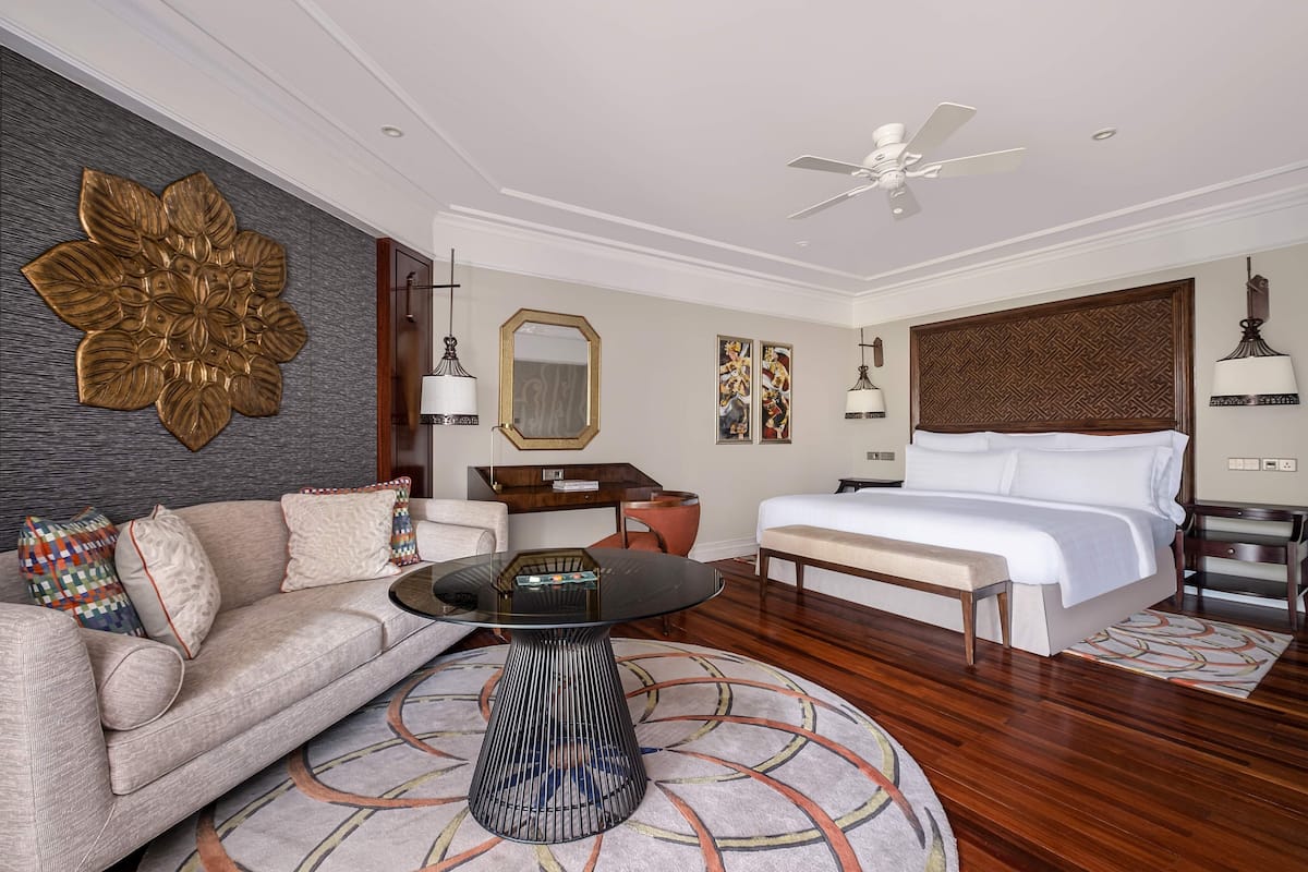 A hotel room with a large bed, a sofa, a coffee table, a ceiling fan, and a decorative wall piece.