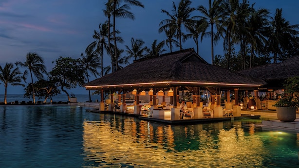 A waterfront bar with a thatched roof, overlooking a pool and palm trees. - InterContinental Bali Resort by IHG (Jimbaran)
