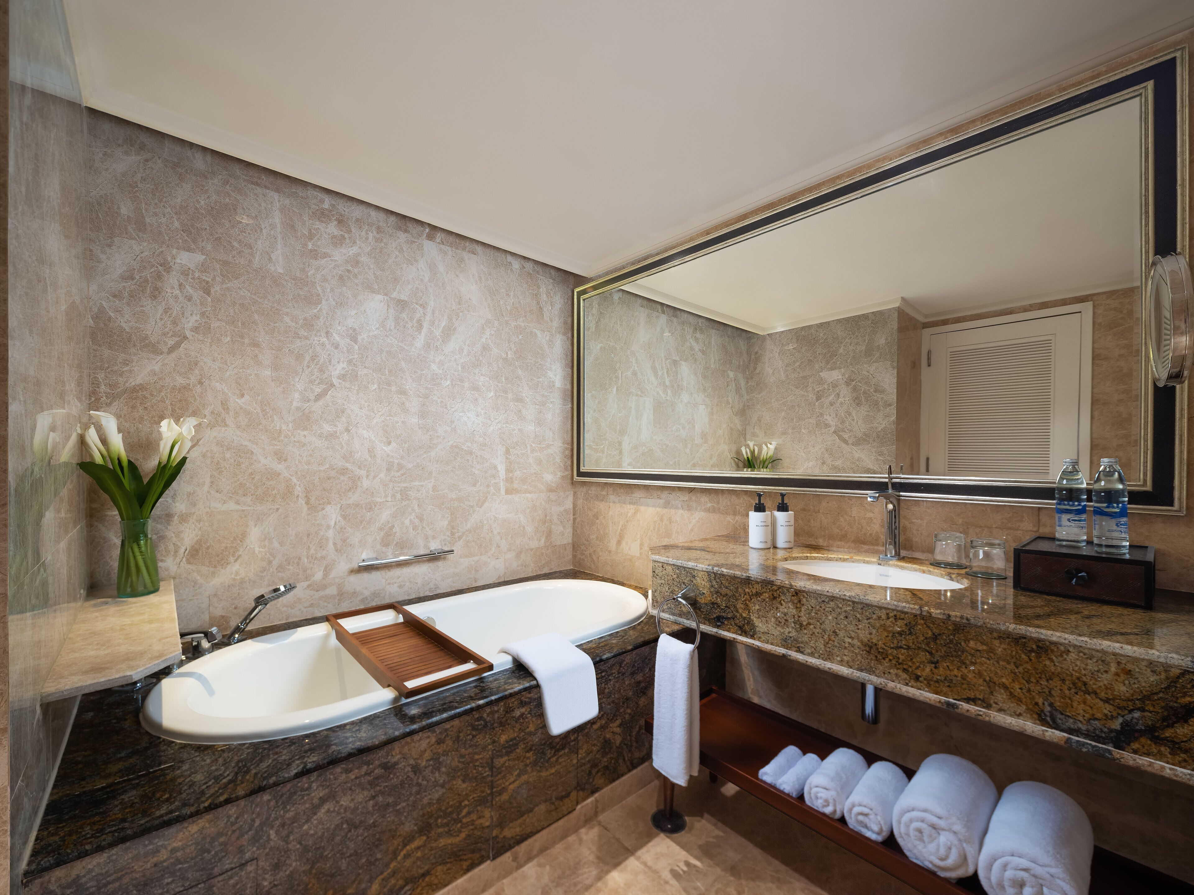 A modern bathroom with a marble wall, a large mirror, a bathtub, a sink, and a towel rack with towels.