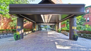 Un passage couvert mène à l'entrée d'un bâtiment dont la façade est en briques, agrémentée de plantes en pot et offrant une vue dégagée sur la rue.