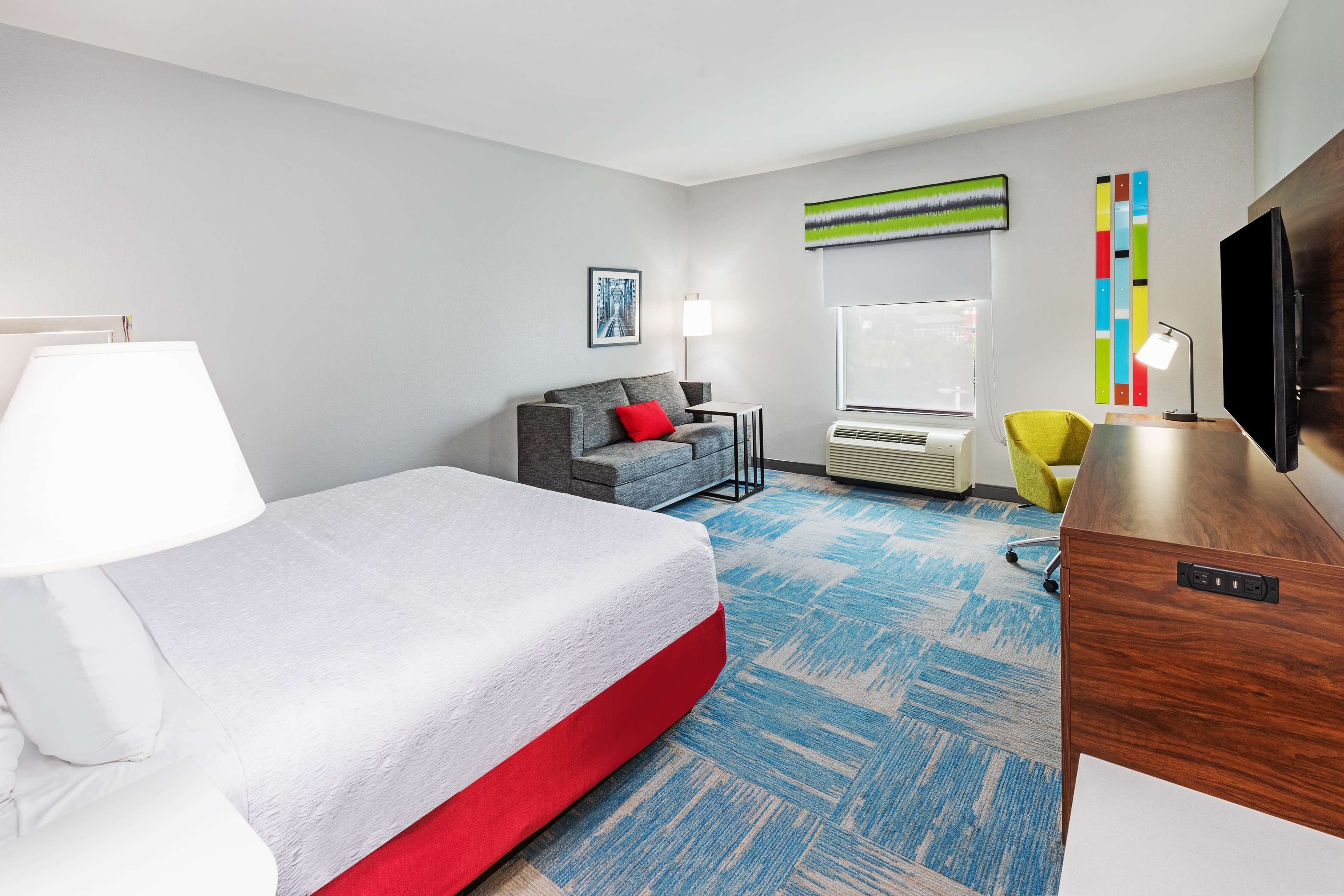 Habitación de hotel con cama, escritorio, silla, televisor, lámpara y ventana.