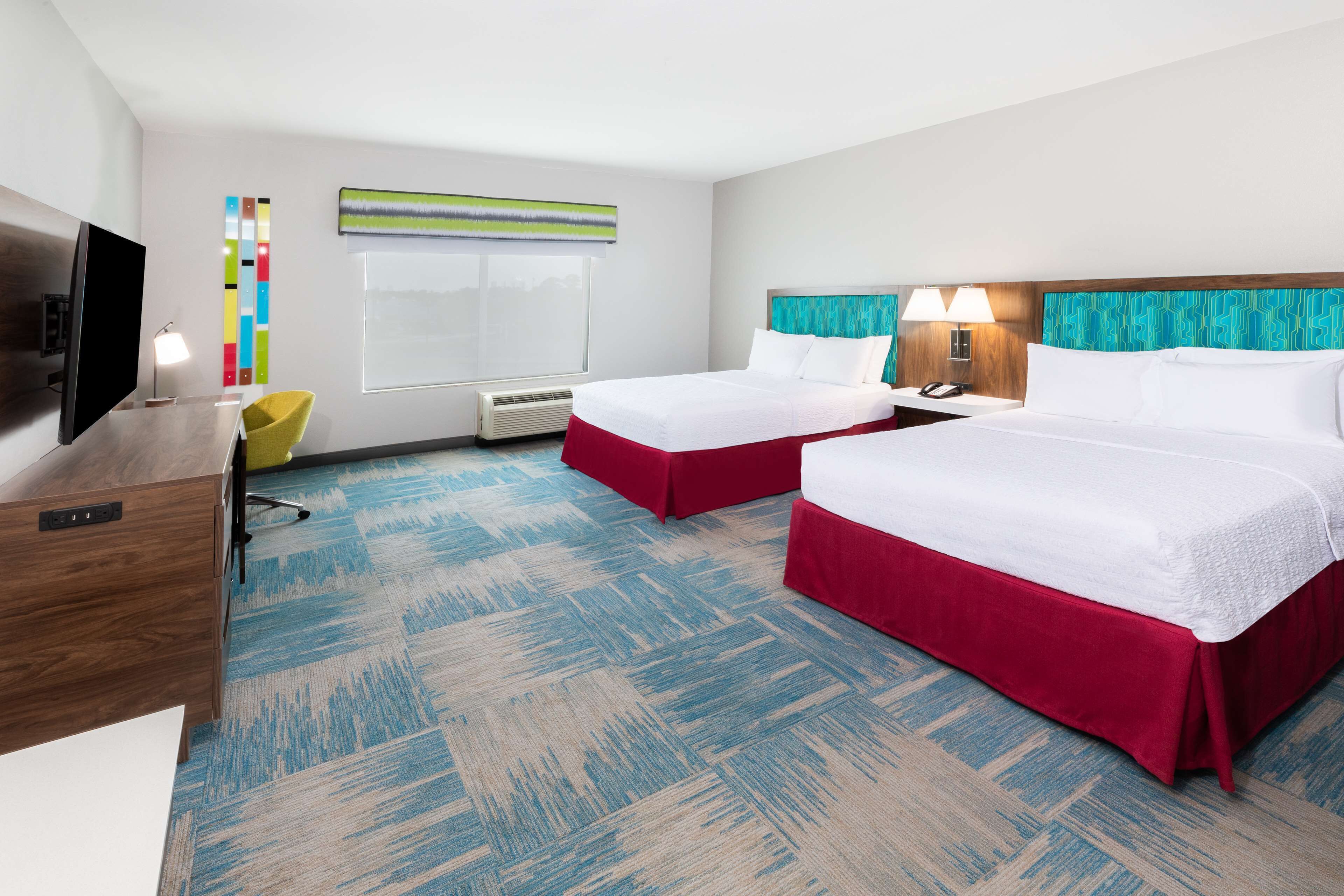 A hotel room with two beds, a desk, a TV, a lamp, and a window with blinds.