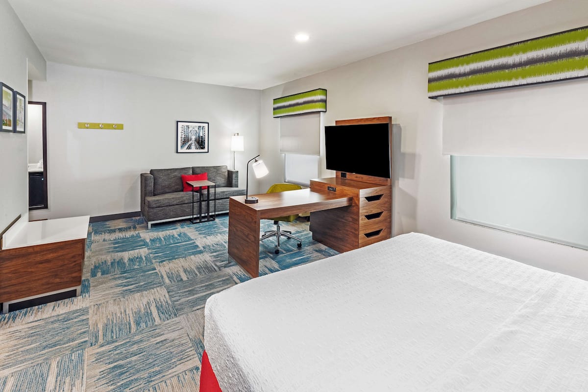 A modern hotel room with a bed, a desk, a chair, a sofa, a TV, and a window.