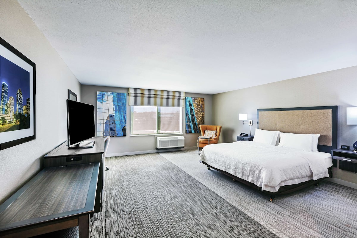 A hotel room with a bed, a desk, a TV, a window with curtains, and a painting on the wall.