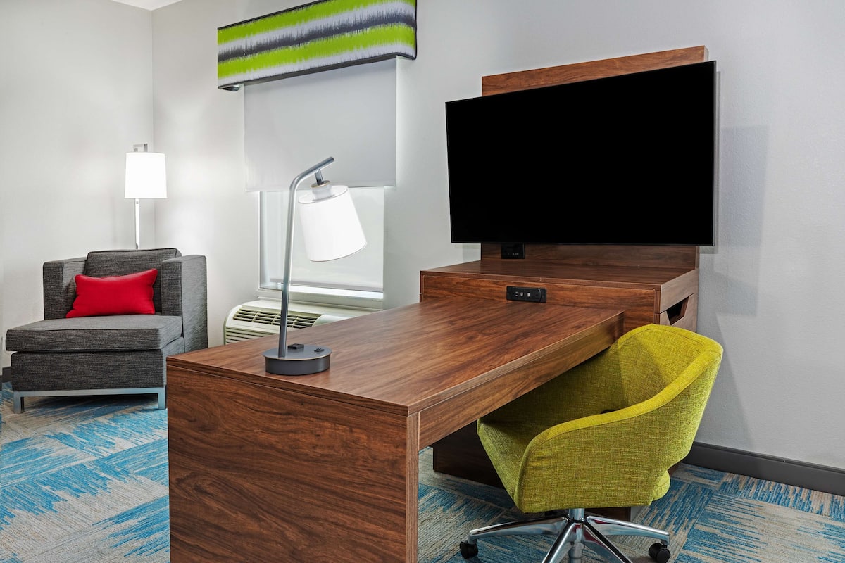A modern office space with a wooden desk, a grey office chair with a red cushion, a yellow office chair, a flat-screen TV, and a lamp.