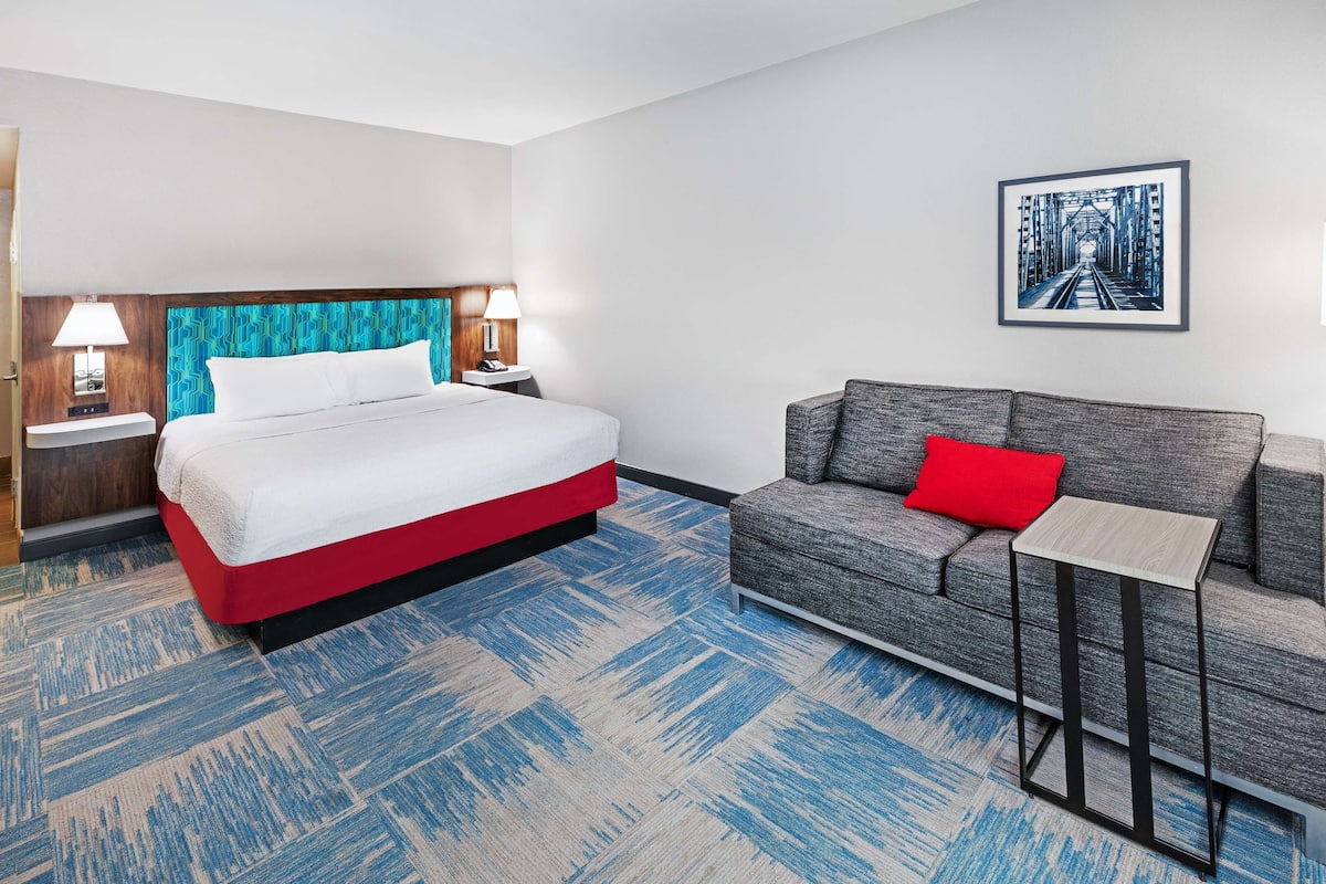 A hotel room with a bed, a gray sofa with a red pillow, a small table, and a framed picture on the wall.