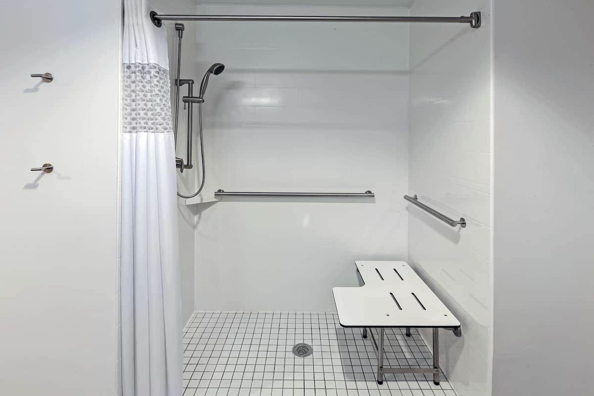 A shower area with a white tiled floor, a showerhead, a grab bar, and a small table.