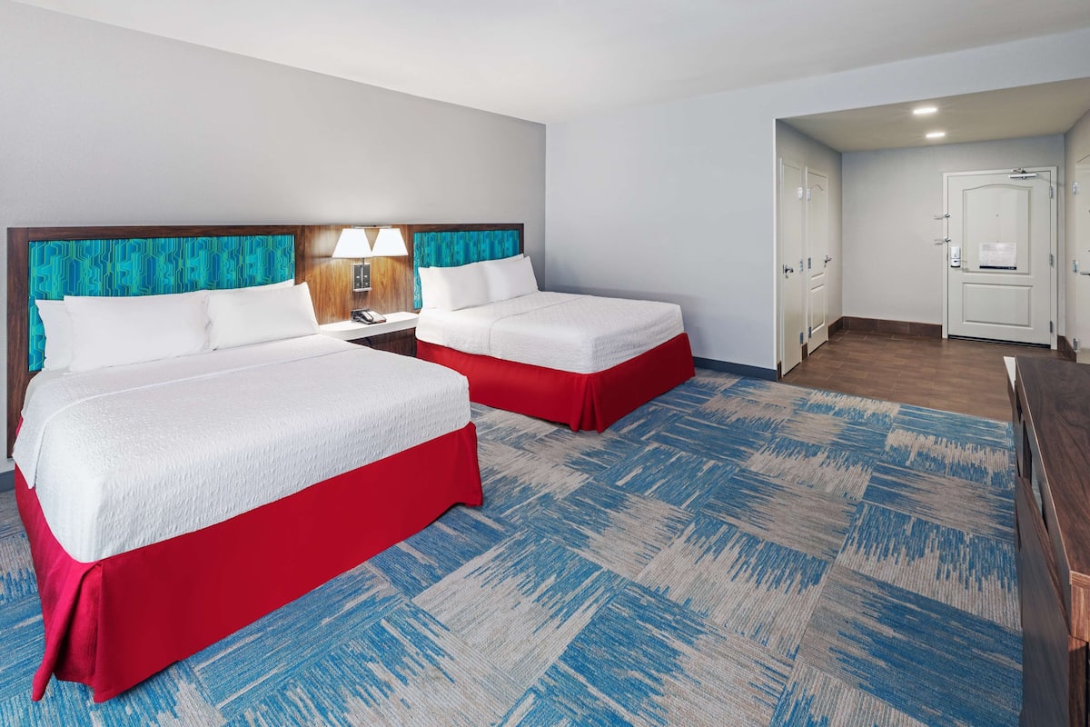 A hotel room with two beds, a desk, a lamp, and a door.