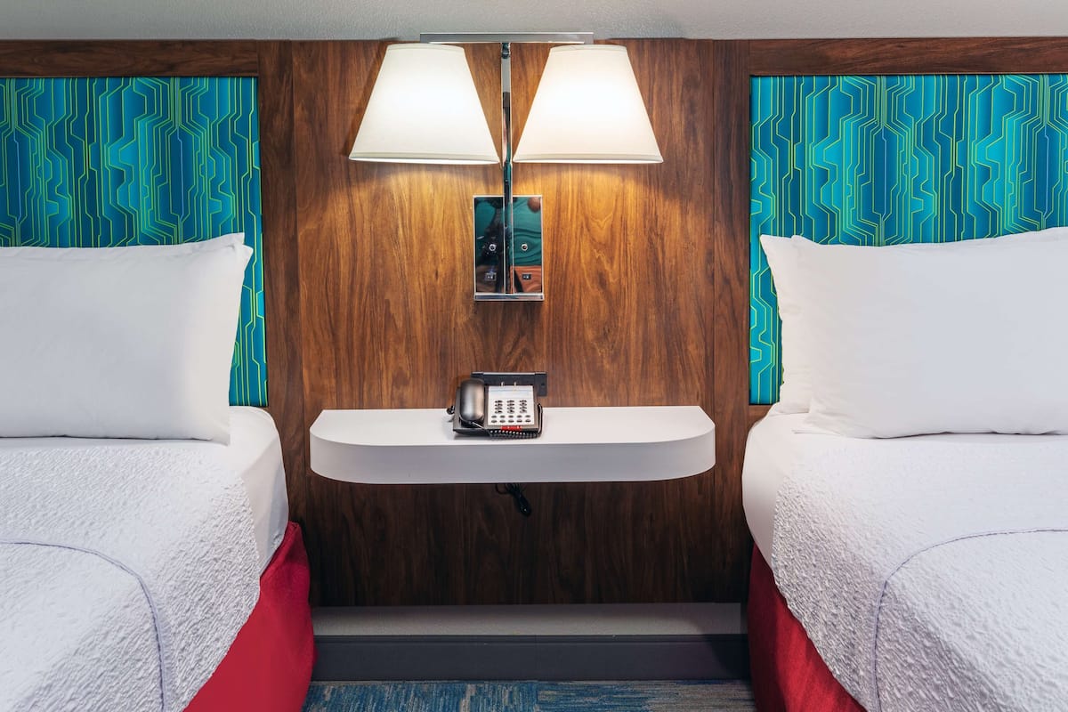 A hotel room with two beds, a bedside table with a phone, and a wall-mounted lamp.