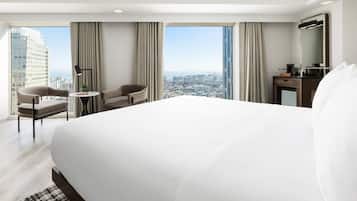 A hotel room with a large bed, two armchairs, a small table, a desk with a mirror, and a view of the cityscape.