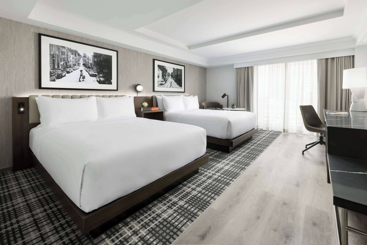 A hotel room with two beds, a desk, a chair, and a large window with curtains.