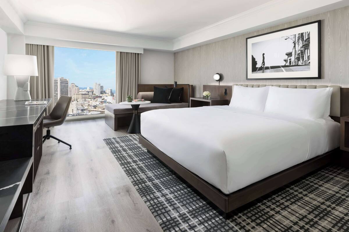 A hotel room with a large bed, a desk, a chair, a TV, and a view of the city.