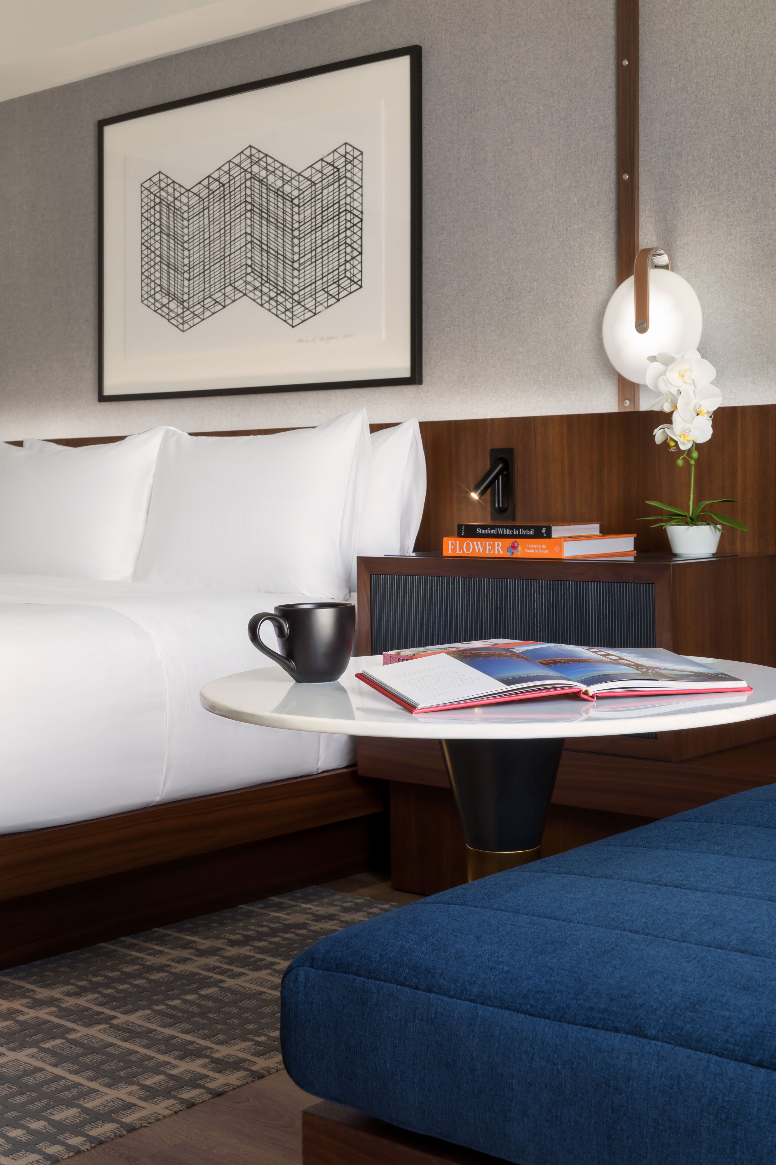 A hotel room with a bed, a bedside table with a lamp, a framed artwork, and a small potted plant.