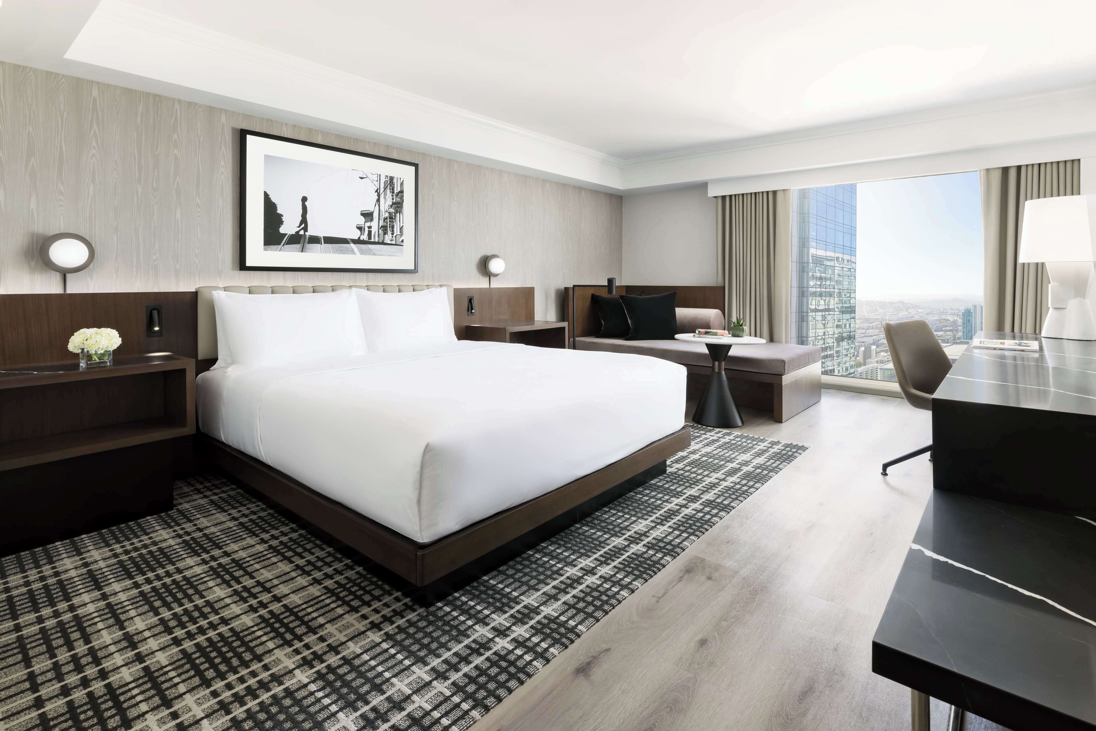 A modern hotel room with a large bed, a desk, a chair, and a view of the city.