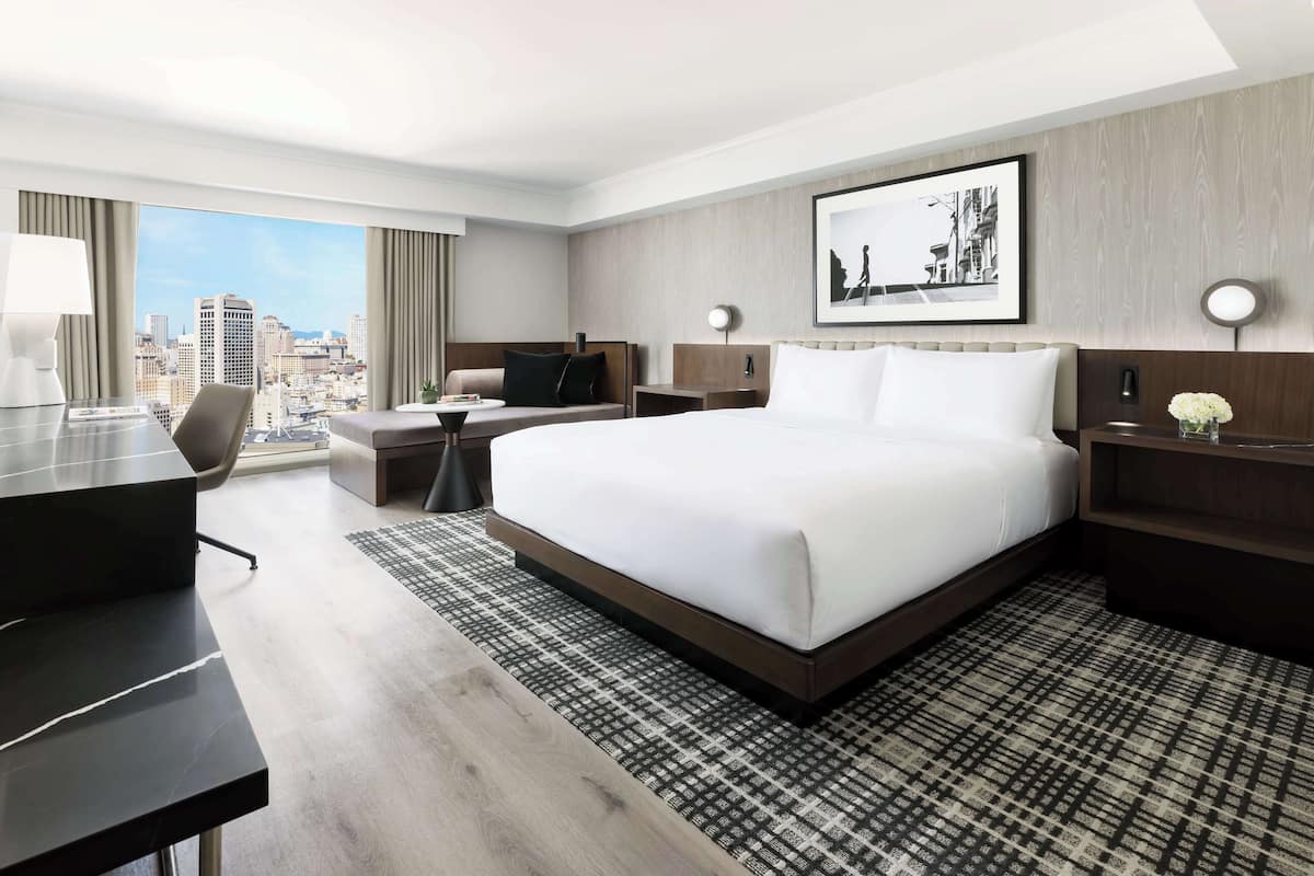 A hotel room with a large bed, a desk, a chair, a small table, and a view of the cityscape.