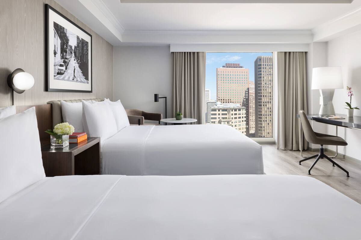 A hotel room with two beds, a desk, a chair, a small table, and a large window with a city view.