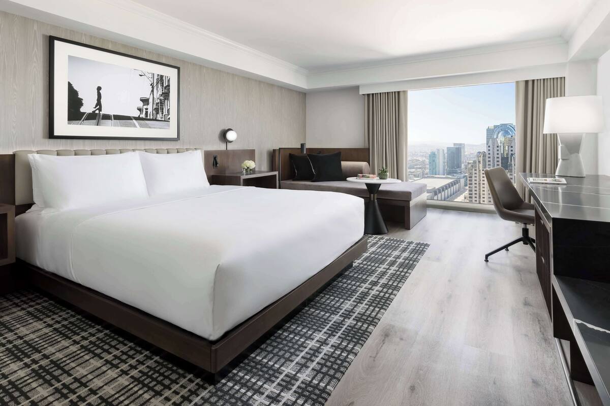 A hotel room with a large bed, a desk, a chair, a small table, a lamp, and a city view.