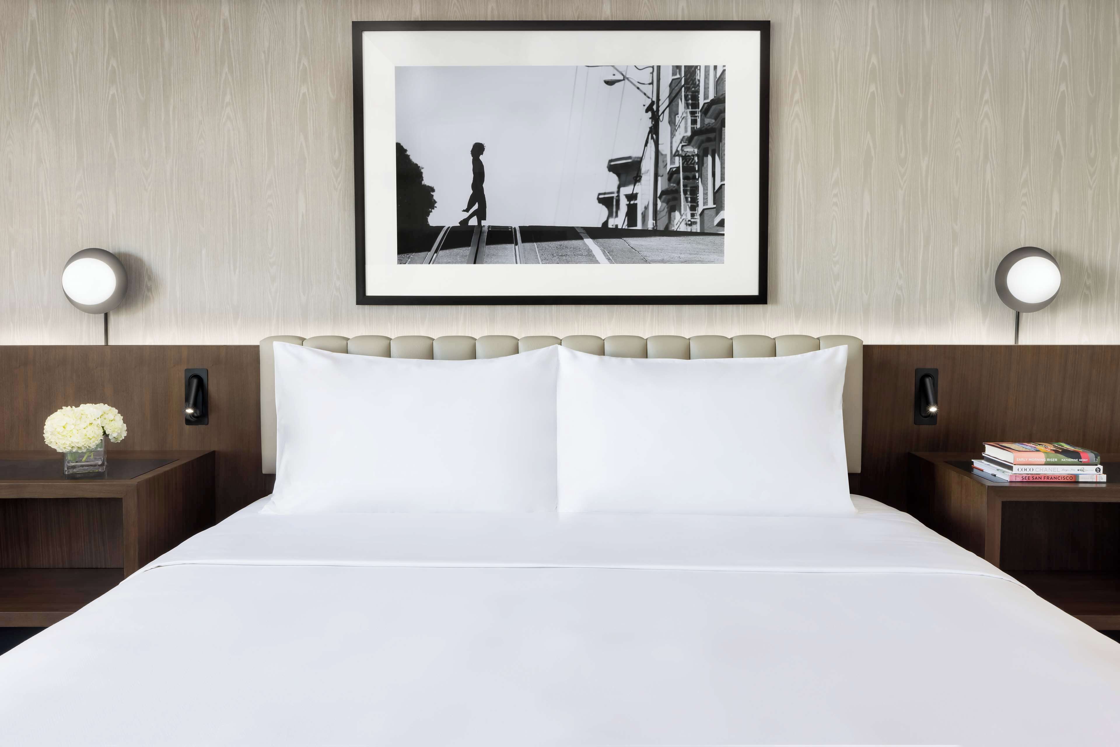 Ein Hotelzimmer mit einem Bett, zwei Nachttischen, einem gerahmten Bild an der Wand und einem Beistelltisch mit Büchern.