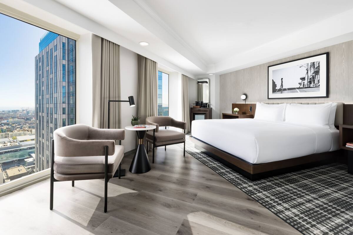 A modern hotel room with a large bed, two armchairs, a small table, and a view of a cityscape through large windows.