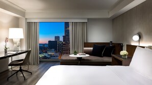 A hotel room with a large bed, a desk, a chair, a small table with a plant, and a view of a cityscape at dusk. - Hyatt Regency San Francisco Downtown SOMA    (San Francisco)