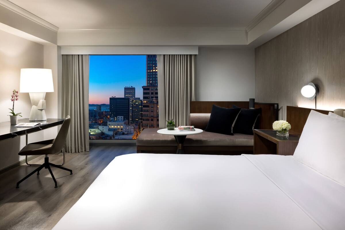 A hotel room with a large bed, a desk, a chair, a small table with a plant, and a view of a cityscape at dusk.