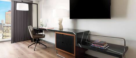 A modern hotel room with a large window, a flat-screen TV, a desk with a chair, and a small bookshelf.