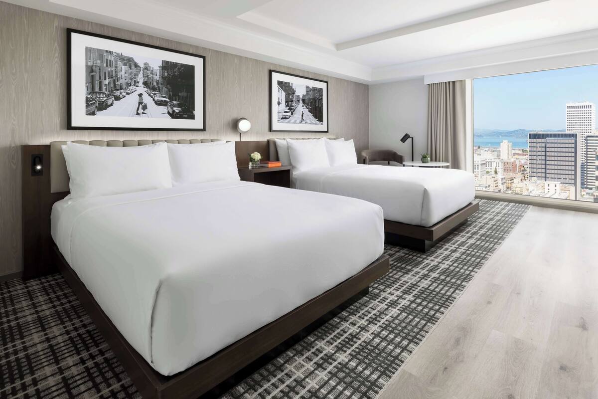 A hotel room with two beds, a large window with a city view, and two framed pictures on the wall.