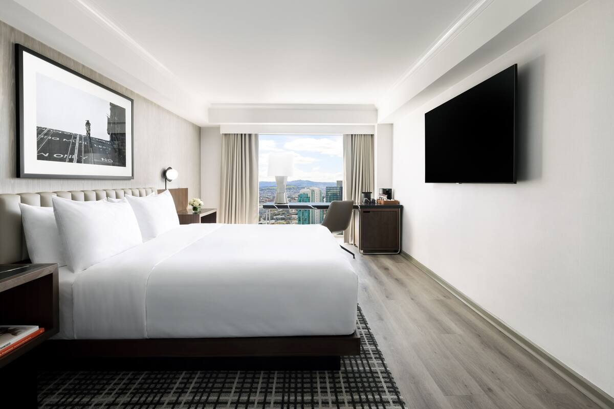 A modern hotel room with a large bed, a flat-screen TV, a desk, and a city view.