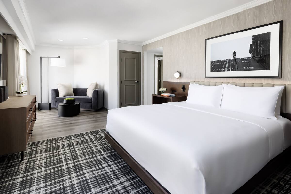 A modern hotel room with a large bed, a sofa, a desk, and a framed picture on the wall.
