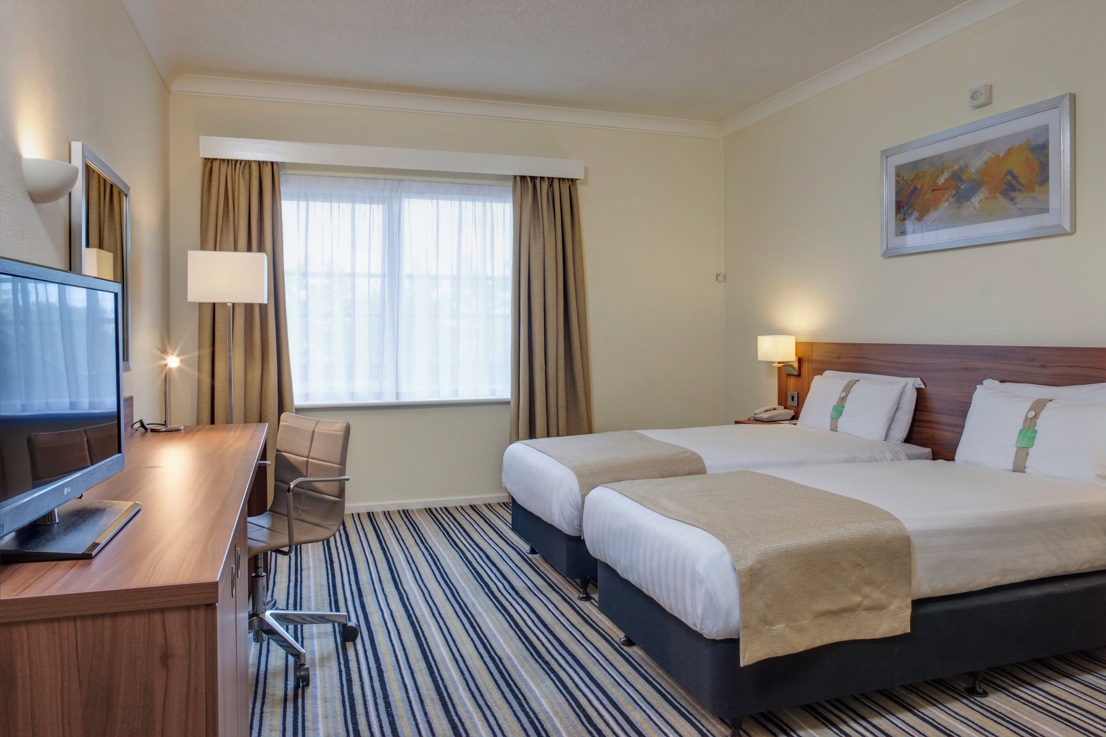 Habitación de hotel con cama, escritorio, silla, televisor, ventana con cortinas y un cuadro en la pared.