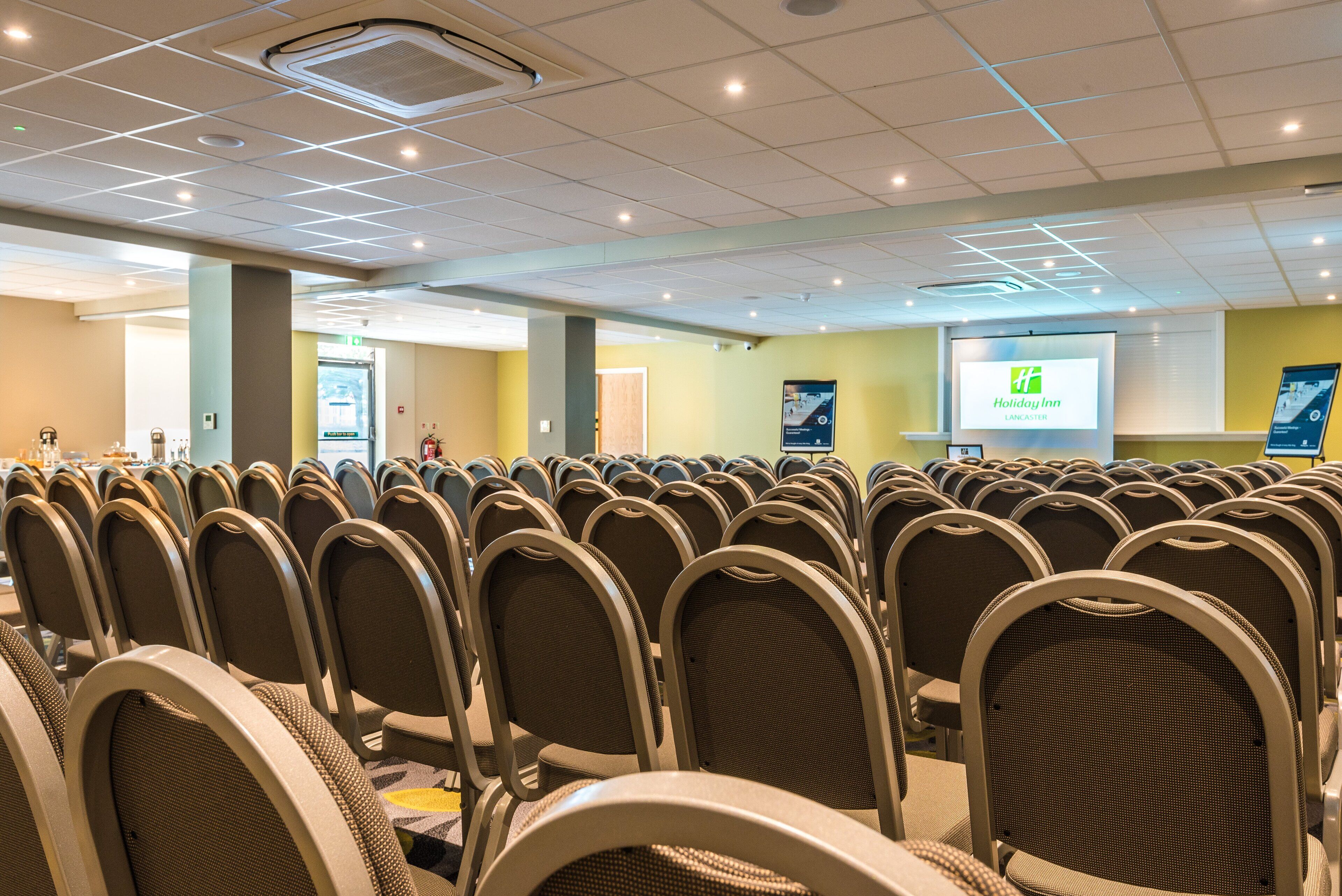 Des rangées de chaises dans une salle de conférence, avec un écran de présentation et une enseigne Holiday Inn.
