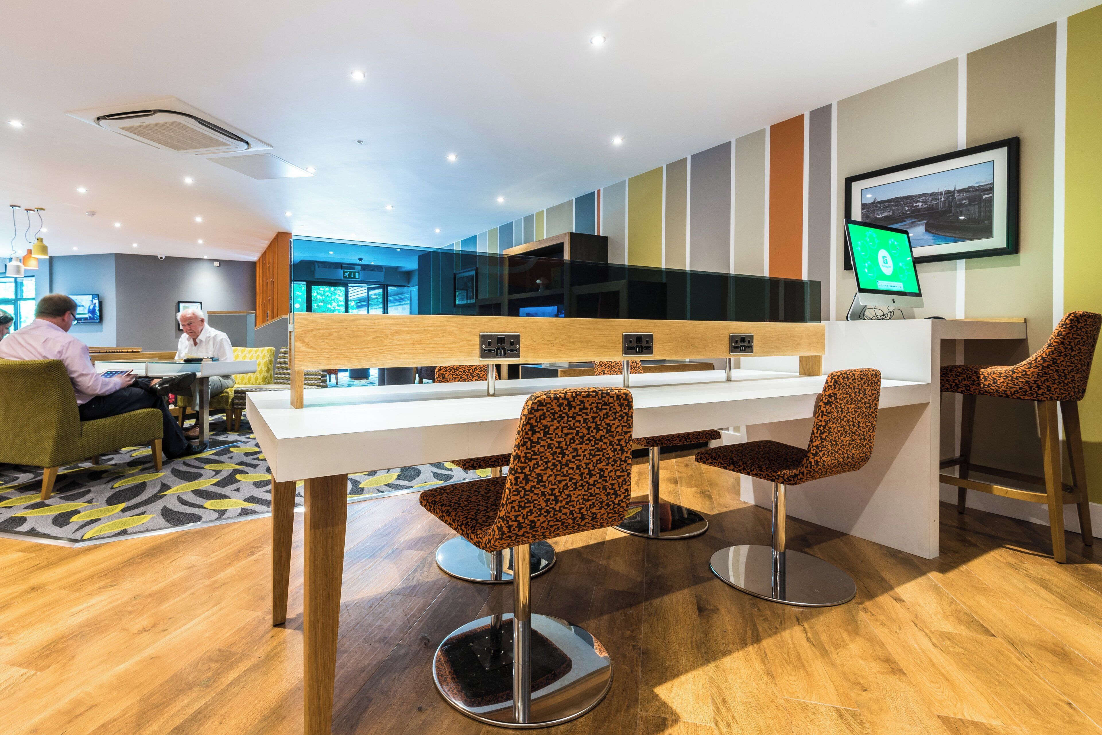 A modern dining area with a long table and patterned chairs, a laptop on a wall, and a view of the interior space.
