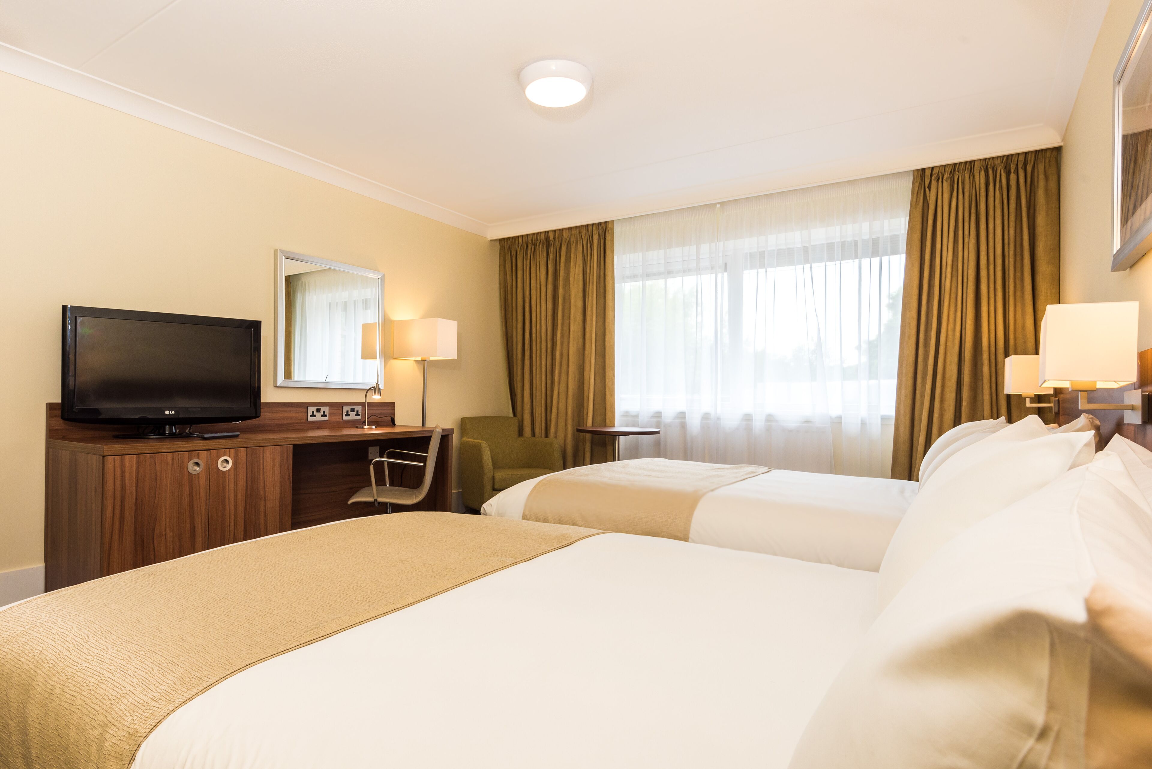 Een hotelkamer met twee bedden, een bureau met televisie, een stoel en een raam met gordijnen.