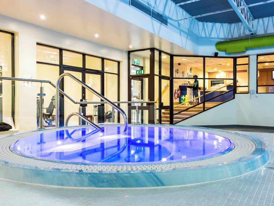 An indoor swimming pool with a glass barrier, a metal staircase, and a visible exit sign.