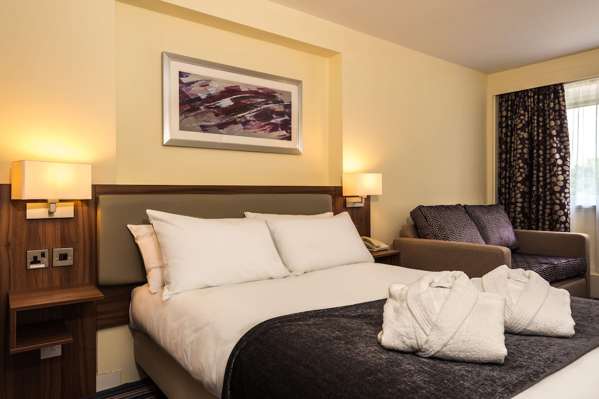 A hotel room with a bed, two bedside lamps, a chair, a painting on the wall, and a window with curtains.