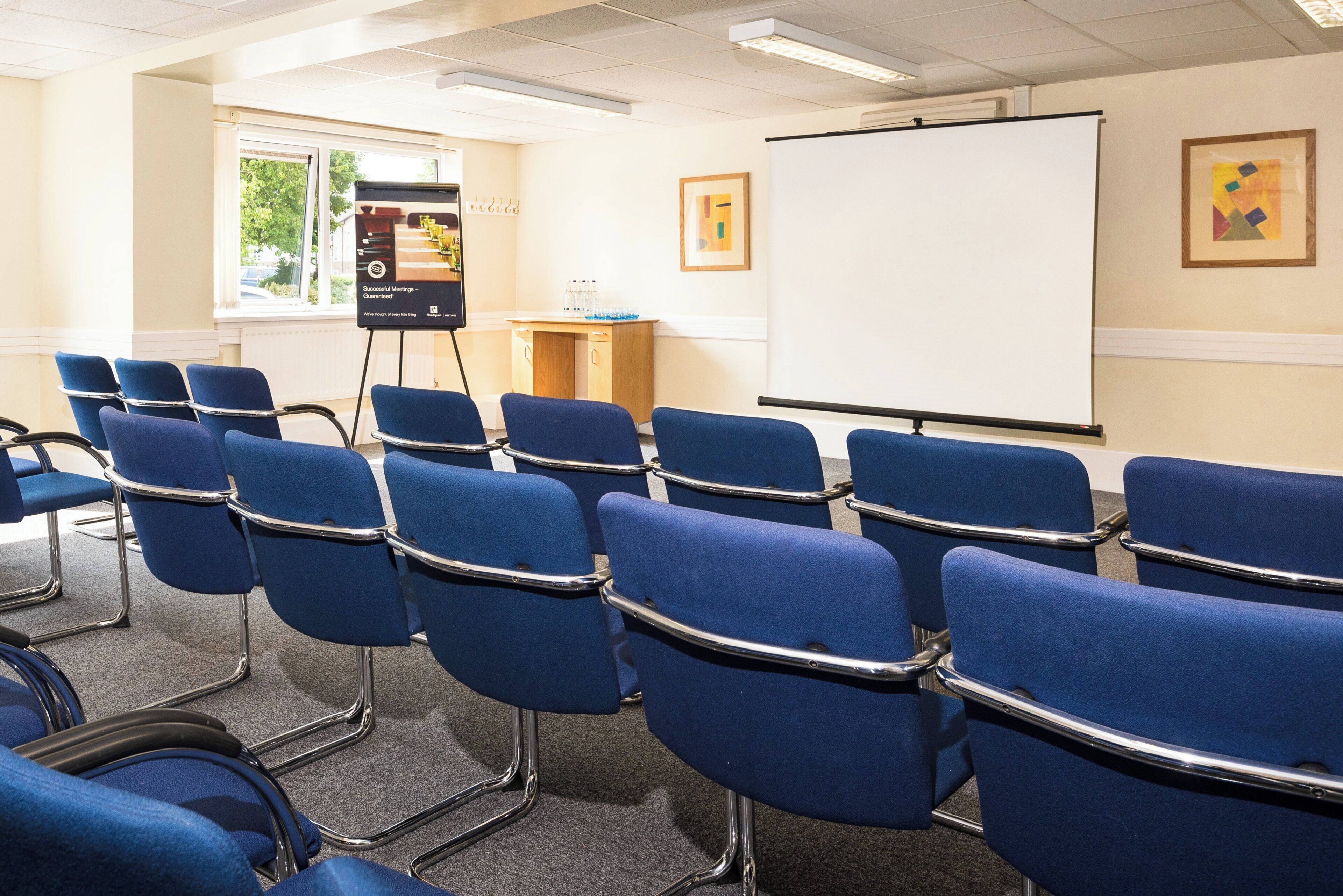 Une salle de conférence avec des rangées de chaises bleues, un tableau blanc et un écran de projection.