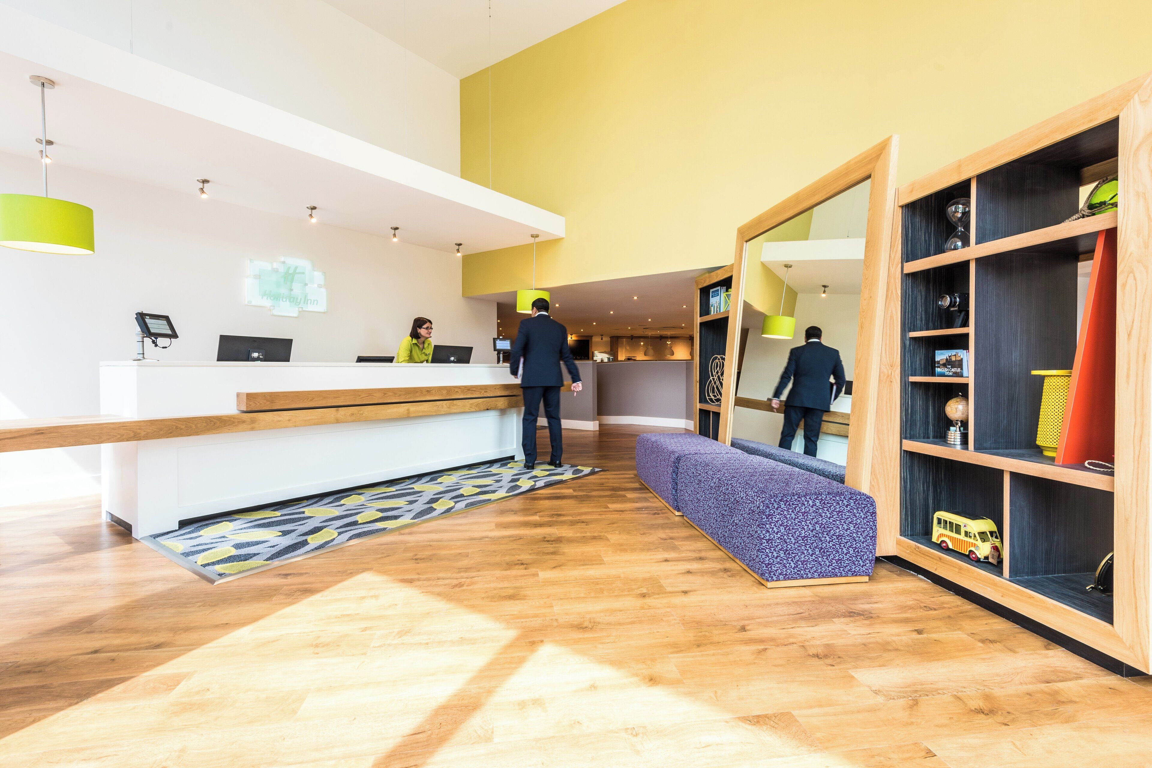 A hotel reception area with a wooden reception desk, a seating area, and a bookshelf with various items.