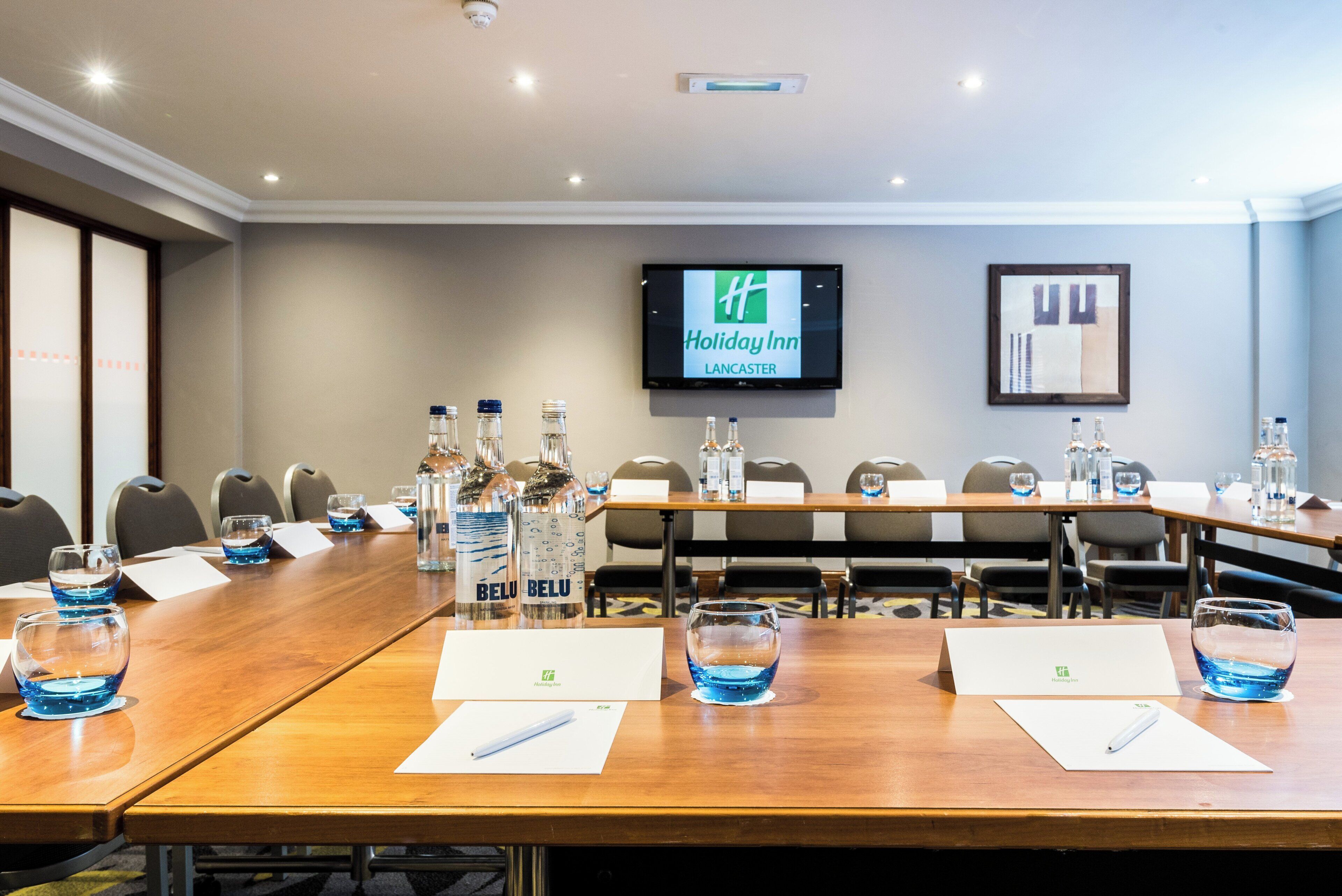 Een vergaderruimte met een lange tafel, stoelen, waterflesjes, glazen en een televisiescherm waarop het Holiday Inn logo staat.