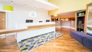 A reception area with a wooden reception desk, a patterned rug, and a seating area with a purple upholstered bench. - Holiday Inn Lancaster by IHG (Lancaster)