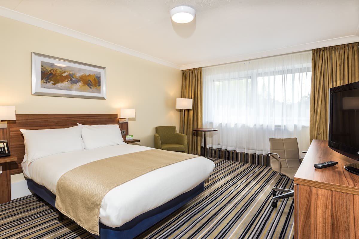 A hotel room with a large bed, a desk with a TV, a chair, a lamp, and a painting on the wall.