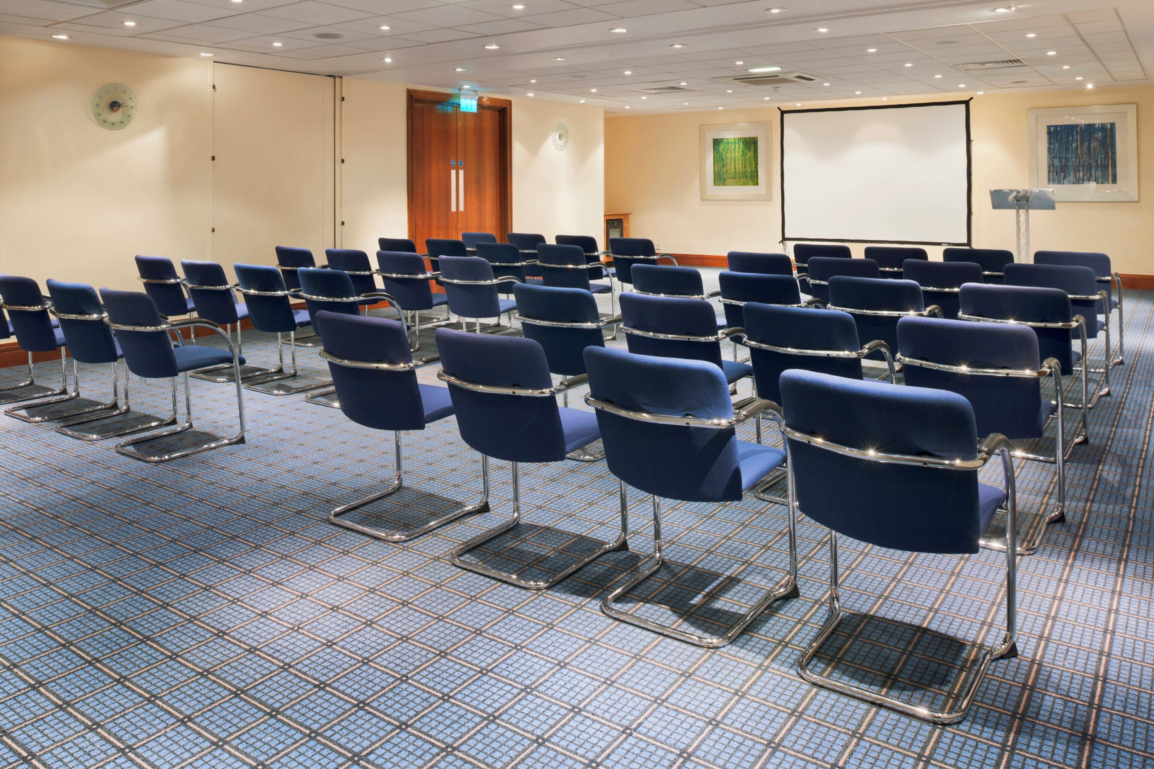 A conference room with rows of blue chairs, a projector screen, and a clock on the wall.
