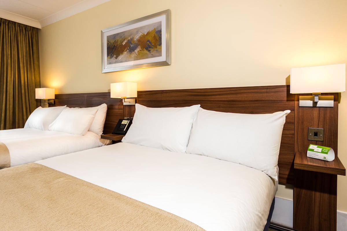 A hotel room with two beds, each with white linens and a beige throw blanket. There are two bedside tables with lamps, a painting on the wall, and a telephone on one of the tables.